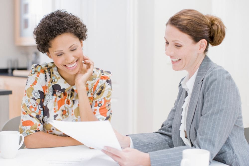 Two women sitting at a table with coffee cups, smiling and looking at a document in a bright office setting, discussing the rehab plans enthusiastically.