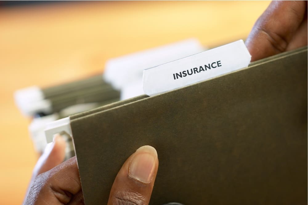 Close-up of a person's hands holding a file folder with a tab labeled "Insurance." Other folders, including one for rehab, are visible in the background.