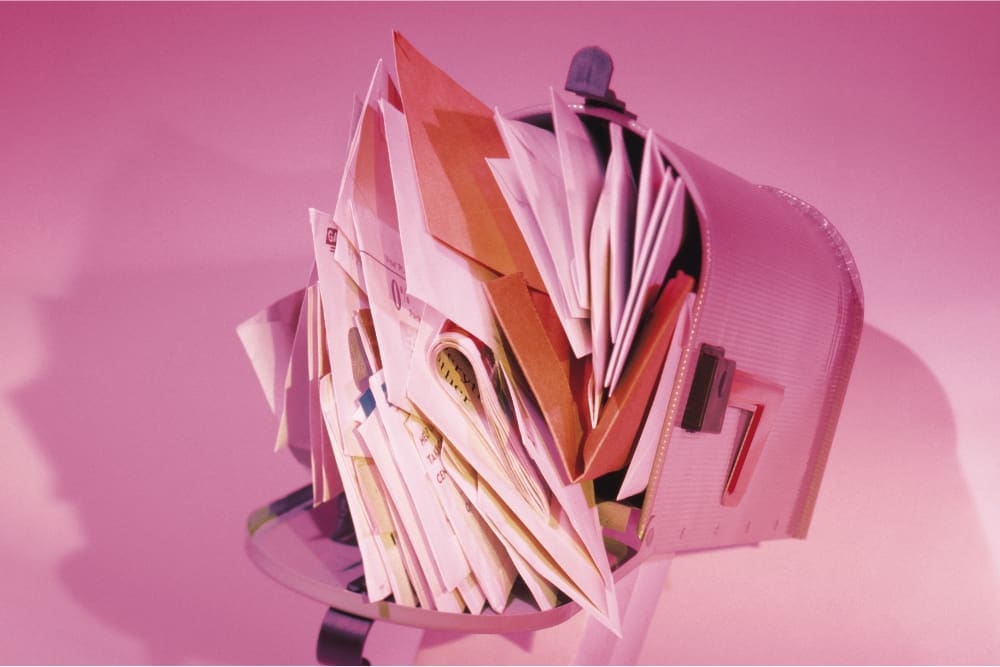 An overstuffed metal mailbox with numerous envelopes and papers spilling out, set against a rehab pink background.