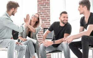 A group of people sitting around a table and enthusiastically giving each other high fives at an addiction recovery center.