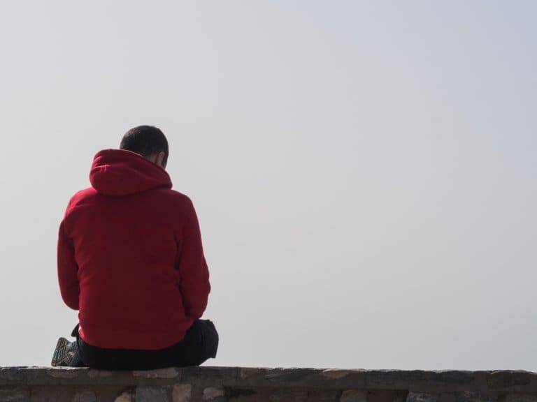 A man in a red hoodie sitting on a wall outside an addiction recovery center.