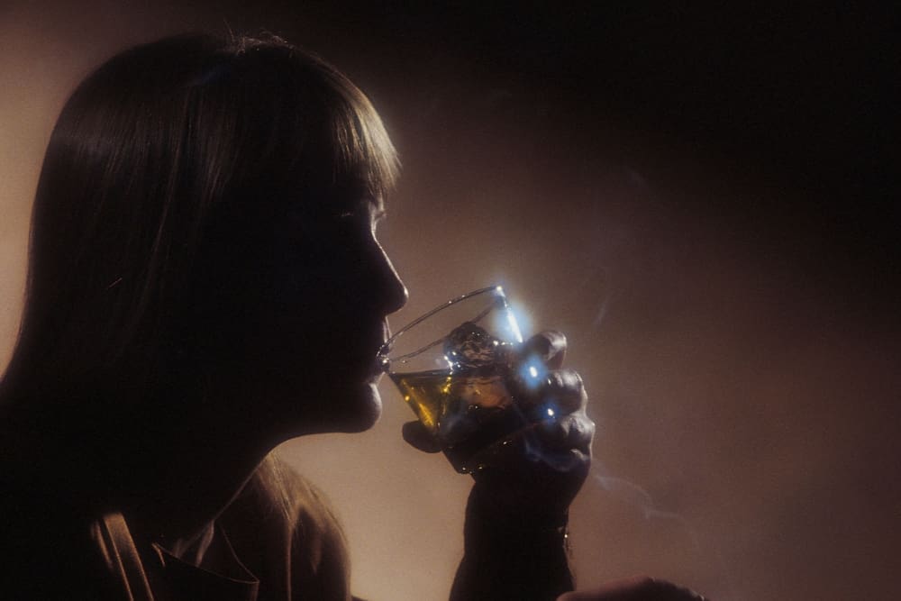 Silhouette of a person with long hair and high eyes drinking from a glass, with a warm backlight creating a moody atmosphere.