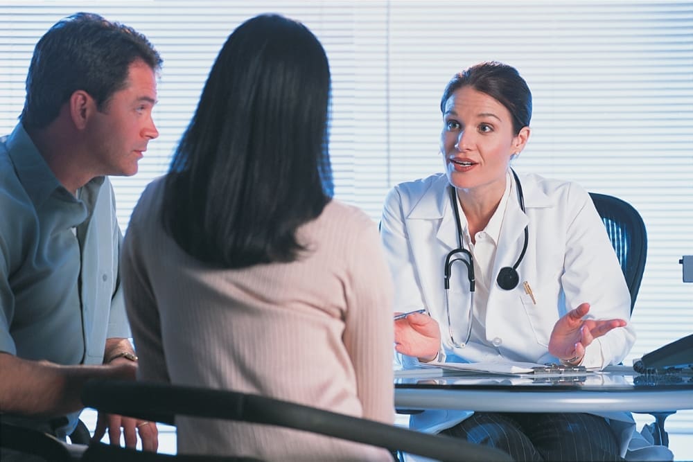 A doctor in a white coat speaks to a man and a woman seated across from her in a medical office, discussing the effects of cocaine nose.