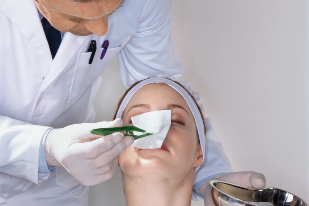 A medical professional in a white coat uses forceps to apply a gauze pad to a patient's face during a procedure, targeting what appears to be remnants of cocaine nose.