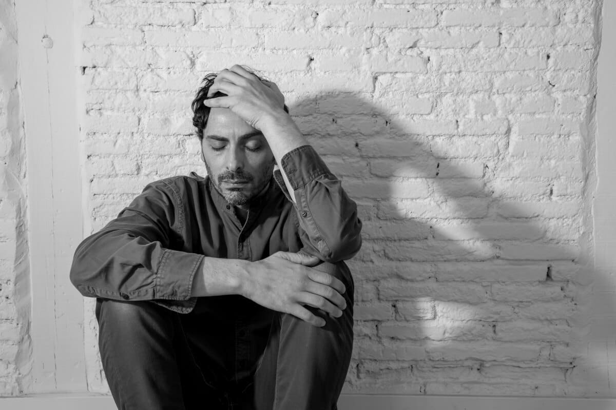 A man sitting against a white brick wall looking down with a hand on his head, appearing contemplative or distressed, possibly reflecting on his journey through alcohol rehab Ohio.