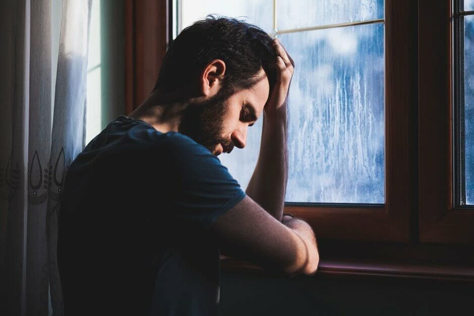 A man resting his head in his hand, looking down thoughtfully by a window with sunlight filtering through at a Drug & Alcohol Rehab center in Ohio.