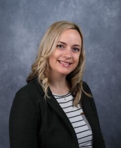 A woman with shoulder-length blonde hair and a black blazer smiles at the camera in front of a grey textured background, aptly representing our polished staff profiles.