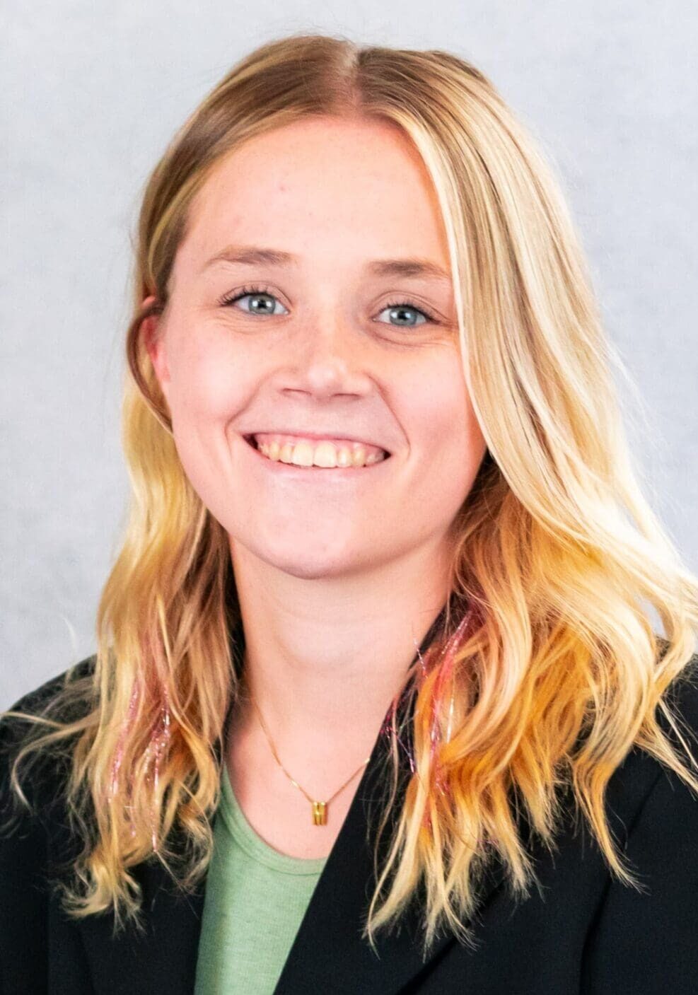 A person with long blond hair, smiling, is wearing a black blazer over a green top in their staff profile. The background is light grey.