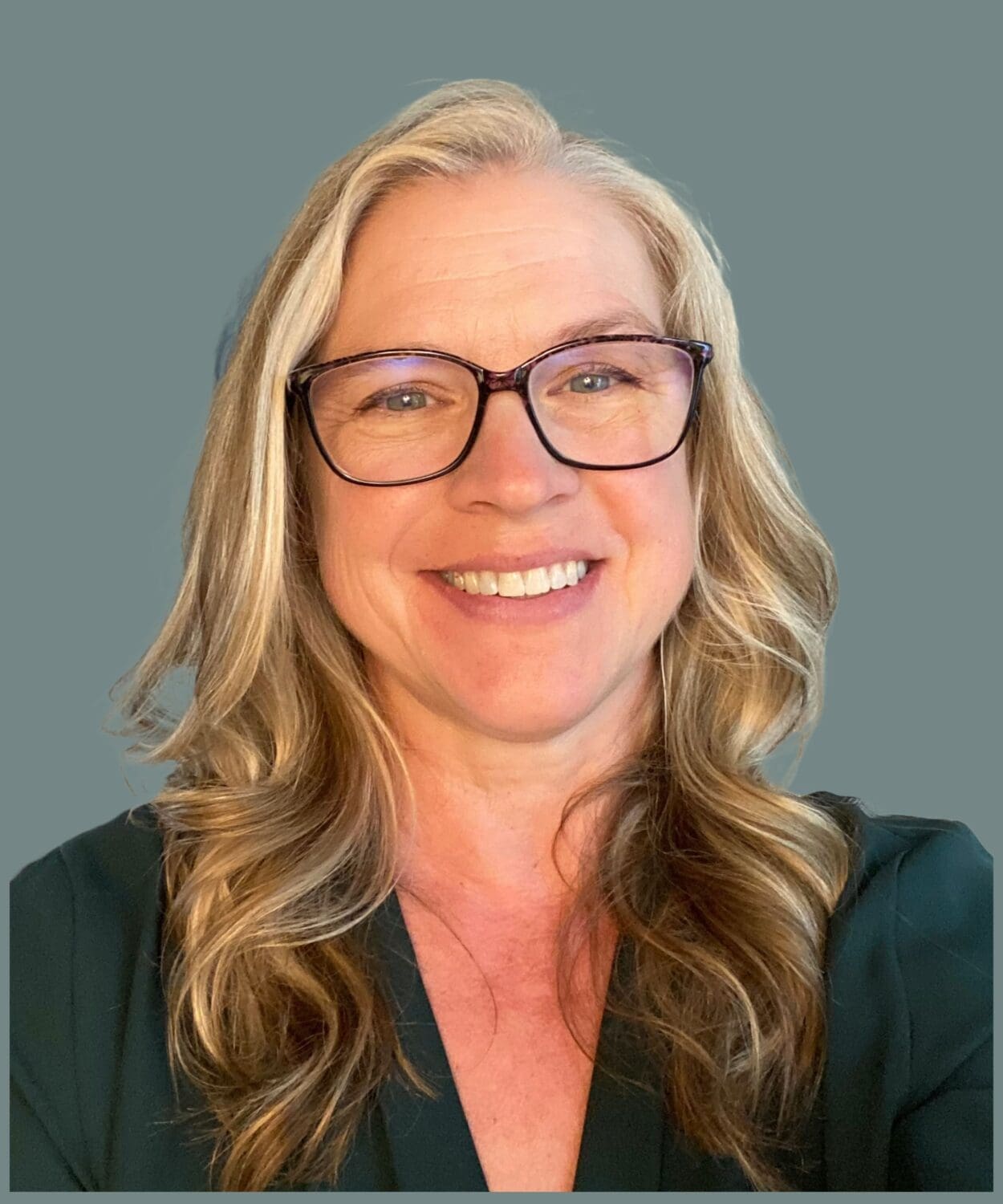 A person with long blonde hair, wearing glasses and a dark green top, is smiling against a plain gray background, perfect for staff profiles.