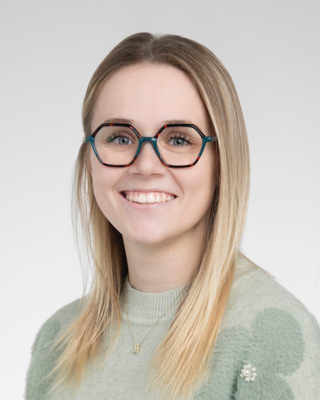 A woman with long blonde hair and octagonal glasses, featured in our staff profiles, wears a light green sweater and smiles warmly against a plain background.